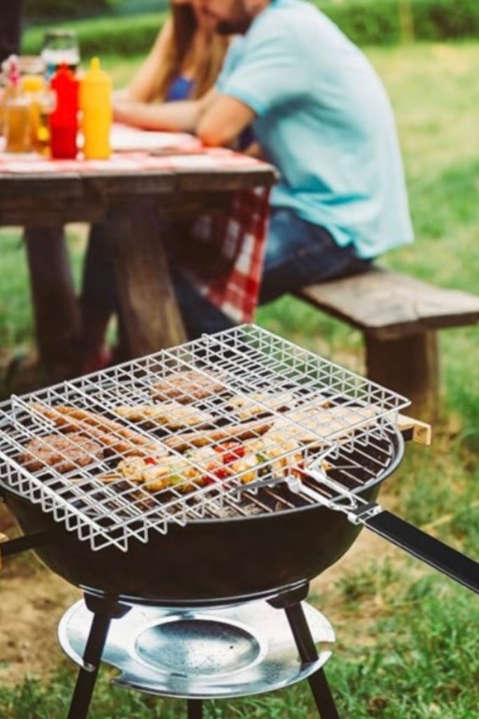 A stainless steel wire grill basket with a long handle being used on a kettle grill to cook fish and vegetables outdoors.