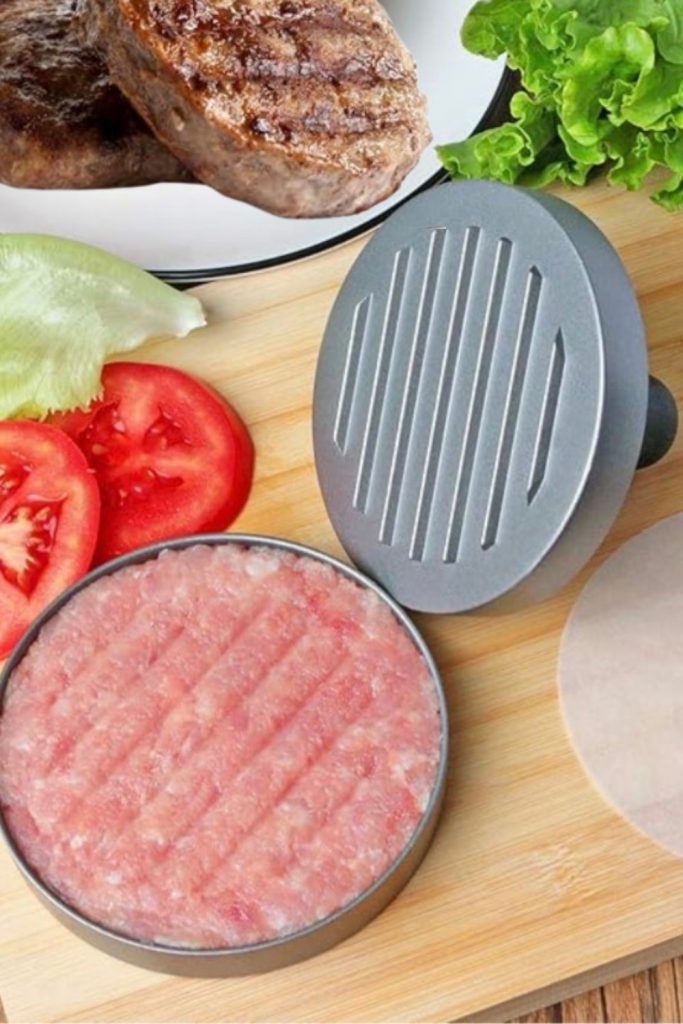 A close-up of a grey plastic burger press kit on a wooden cutting board, shown with freshly shaped, uniform homemade meat patties and vegetables.