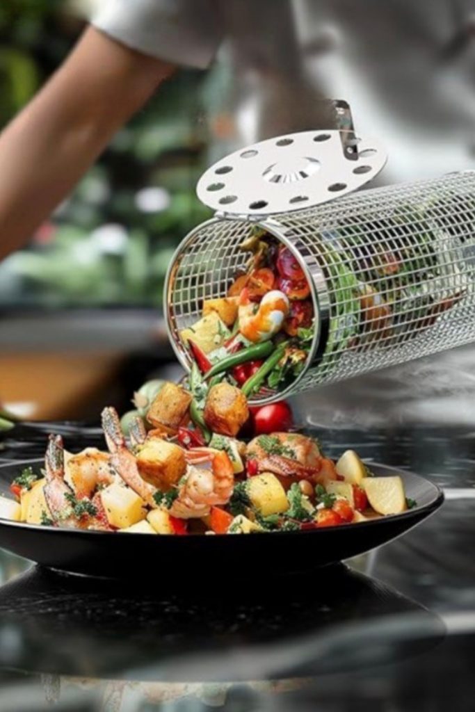 A close-up view of a stainless steel wire mesh rolling grill basket overflowing with roasted chopped vegetables including peppers, onions, and squash.