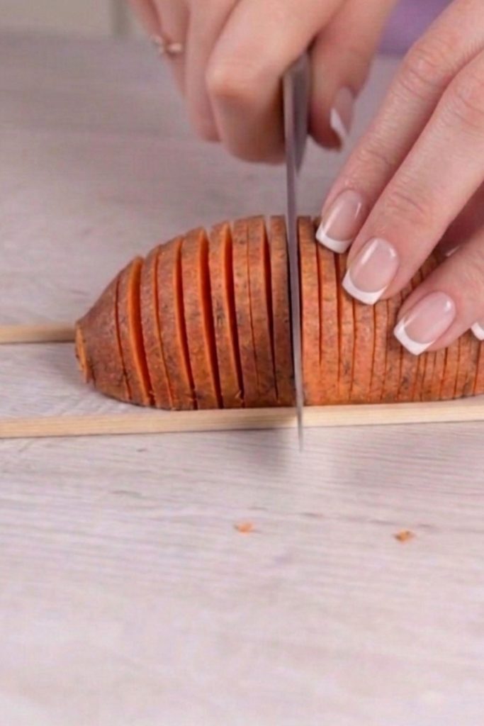 A close-up shot showing the Hasselback cutting technique: a hand slicing a potato placed between two wooden chopsticks to ensure even, thin slices without cutting all the way through. Minimalist and clean kitchen aesthetic.