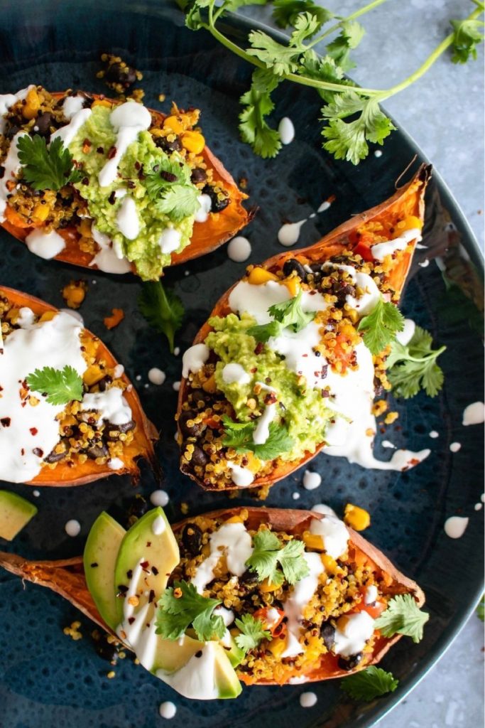 Close-up of a Mexican quinoa stuffed sweet potato garnished with fresh avocado, red peppers, and cilantro, served on a rustic dark plate with lime wedges.