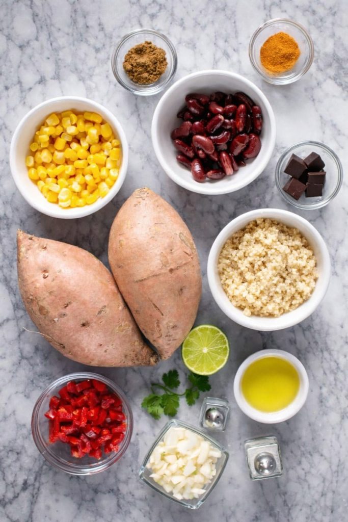 Top-down flat lay view of ingredients for Mexican Quinoa Stuffed Sweet Potatoes, including raw sweet potatoes, quinoa, black beans, corn, peppers, spices, lime, and olive oil in white bowls on a marble surface.